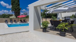 Ett område vid poolen med en pergola, solstolar och en grillplats.