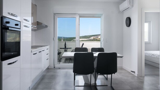 A modern kitchen with white cabinets, a built-in oven, and a dining area with black chairs and a table. - Beautiful apartment for 5 people with A/C, WIFI, TV, balcony and pets allowed (Solin)