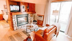 A cozy living room with a stone fireplace, a television mounted on a wooden cabinet, a leather sofa, and a large window with curtains. - Private Forest Retreat | Ruidoso Couples Escape (Ruidoso)