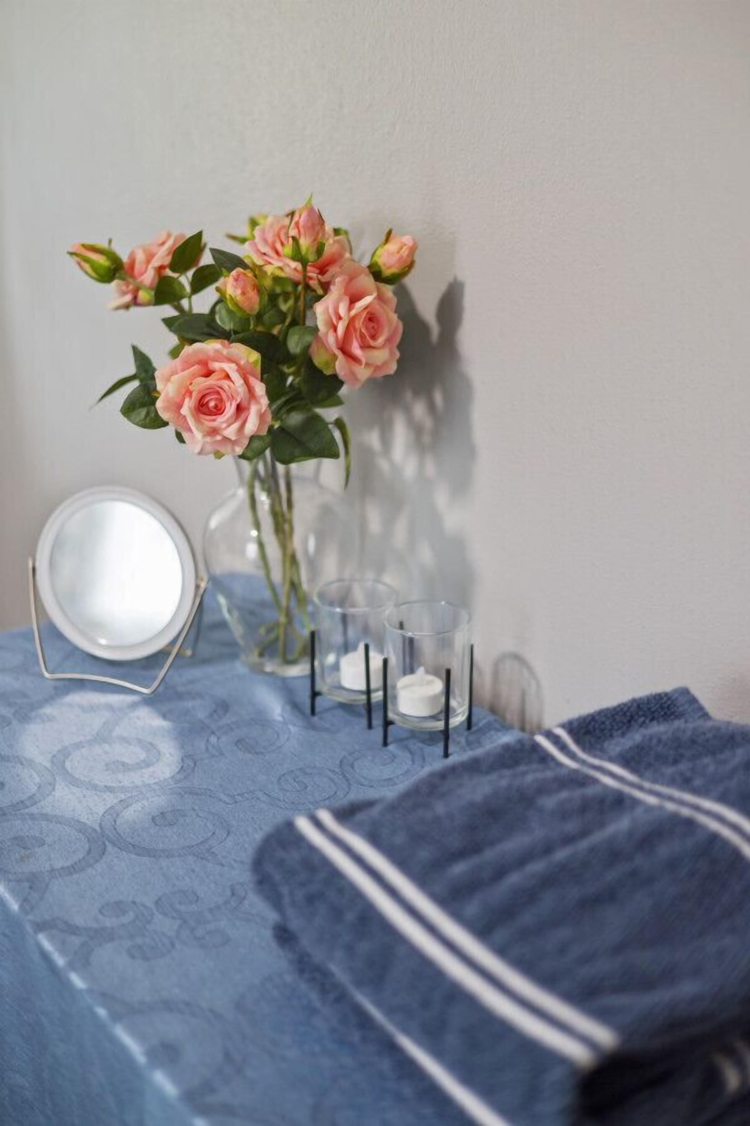 Une salle de bain avec une serviette bleue à motifs, un vase en verre contenant des roses roses et un miroir rond posé sur un support.