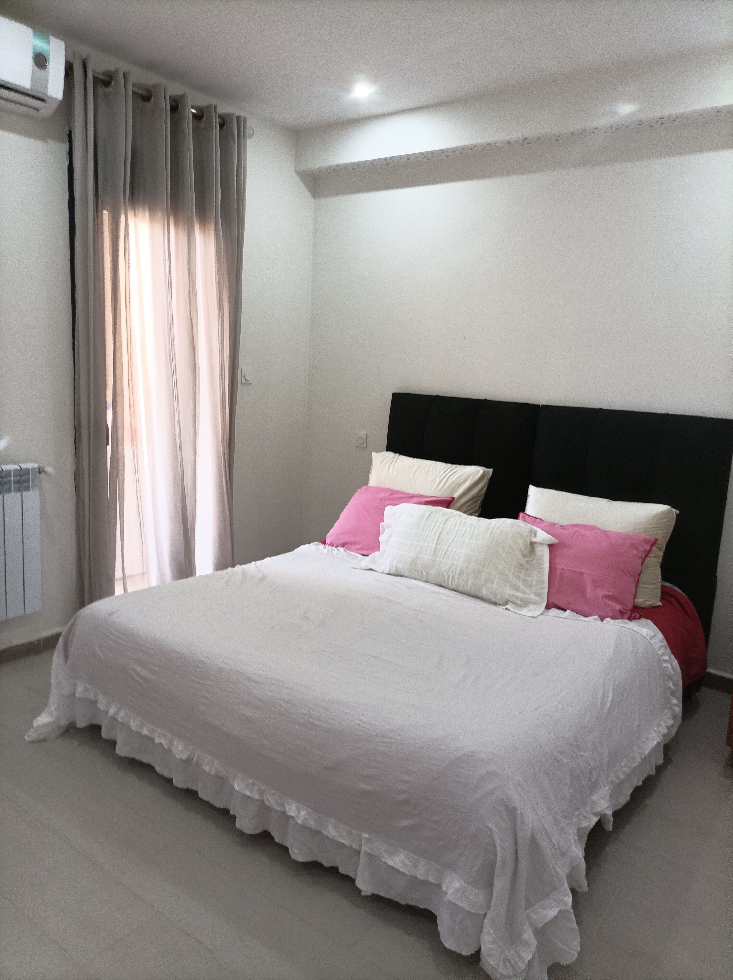 A neatly made bed with white and pink bedding, a black headboard, and a window with curtains.