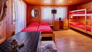 Een knus kamer in chaletstijl met houten wanden en plafond, een bed met rood gestippelde beddengoed, een klein houten bureau, een spiegel en een stapelbed met rood gestippelde beddengoed.