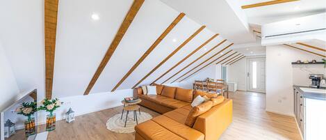 A modern living room with a large sectional sofa, wooden beams on the ceiling, and a kitchen area visible in the background.