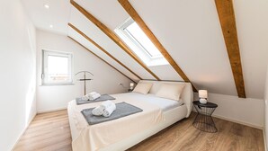 A bedroom with a sloped ceiling, wooden beams, a skylight, a bed with pillows, a bedside table with a lamp, and a floor lamp. - Beautiful apartment in Kastel Novi (Kastel Novi)