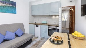 A compact living space with a kitchenette, a sofa with cushions, a dining table with a plate of food, and a bowl of lemons. - Cosy apartment for 4 people with A/C, pool, WIFI, TV and terrace (Tossa De Mar)