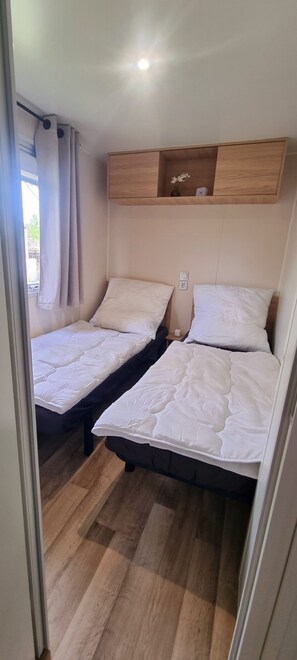 Une chambre à coucher de taille modeste, avec deux lits simples, une étagère en bois au-dessus et une fenêtre avec des rideaux.