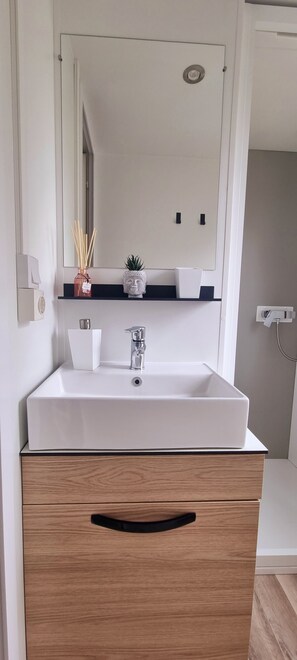 Une salle de bain avec un lavabo blanc, un miroir au-dessus, une petite étagère avec des objets décoratifs et un meuble en bois en dessous.