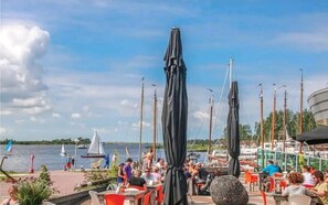 Un restaurant situé au bord de l’eau, avec une terrasse, des voiliers en arrière-plan et un ciel dégagé.