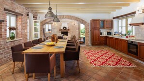 A spacious kitchen with a long dining table, wooden cabinets, and a brick wall.