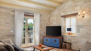 A cozy living room with stone walls, a flat-screen TV on a wooden cabinet, a comfortable sofa, and a coffee table with a book and candle.