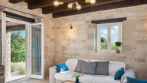 A modern living room with a stone wall, a white sofa with blue and gray cushions, a wooden coffee table, and a window with a view of greenery. - Wonderful private villa with private pool, WIFI, A/C, TV, terrace, pets allowed and panoramic view (Pazin)