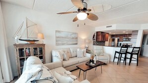 A modern living room with a sofa, coffee table, and a bookshelf. There is a kitchen area with a dining table and chairs visible in the background.
