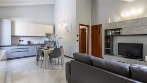 Ein modernes Wohnzimmer mit grauem Sofa, einem Esstisch mit Stühlen, einer Küchenzeile mit Schränken und Geräten sowie einem an der Wand montierten Fernseher.