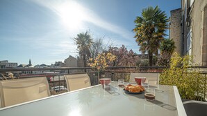 Una mesa con cubierta de vidrio, dos sillas, un plato de croissants y dos tazas de café en un balcón con vistas a un paisaje urbano de edificios y árboles.