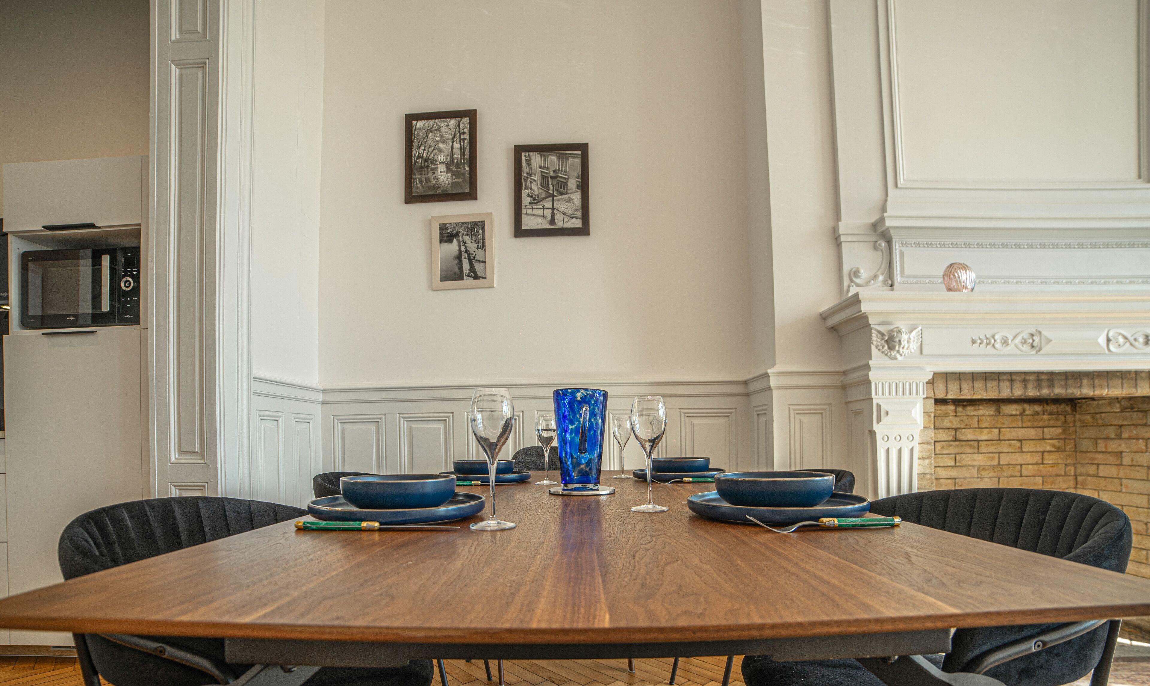 A dining room with a wooden table set for six, surrounded by black chairs. There are three framed black and white photographs on the wall.