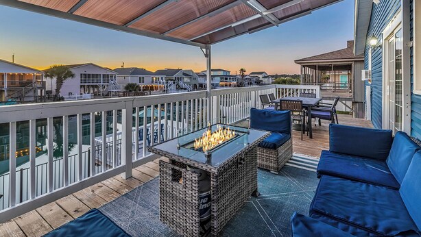 A covered outdoor deck with a glass-top fire pit, wicker furniture, and a view of neighboring houses. - Waterfront Home: 4 min walk to beach, Pool & Mini-Golf (North  Myrtle Beach)