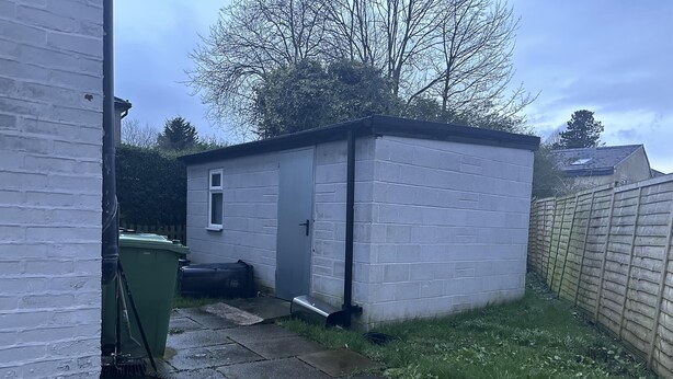 Un petit abri de plain-pied, doté d’une porte et d’une fenêtre, situé à côté d’un mur en briques blanches et d’une clôture en bois.