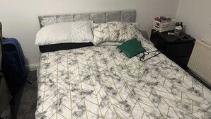 Un lit avec une couvre-lit à motifs de marbre, une tête de lit grise et une table de chevet sur laquelle se trouvent un panier et une bouteille.