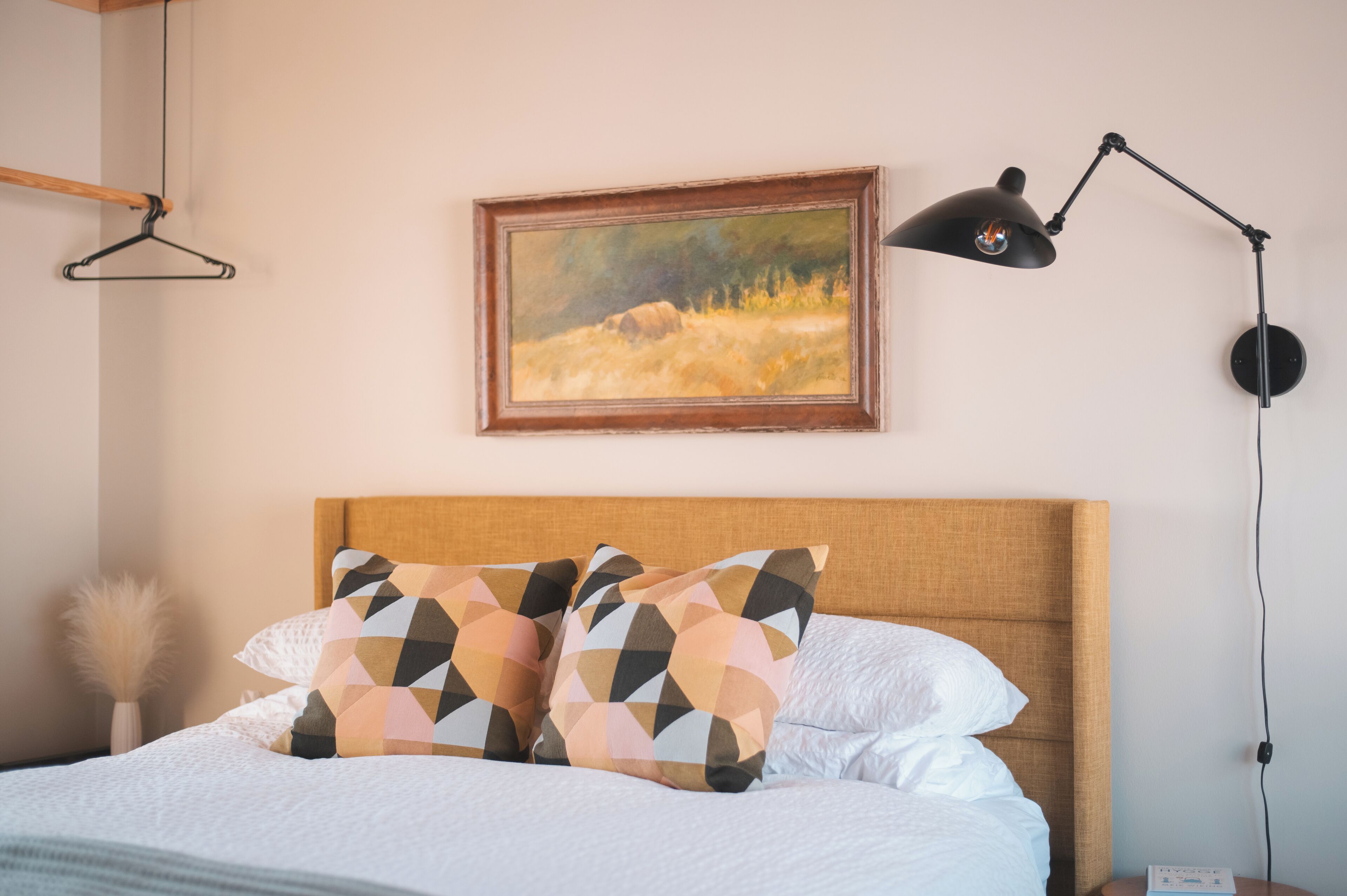 Een net opgemaakt bed met gestreepte kussens, een houten hoofdbord, een schilderij van een landschap en een bureaulamp.