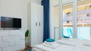 Una habitación de hotel moderna con televisor de pantalla plana, un armario blanco, una cama grande con ropa de cama blanca, una cortina azul y un balcón con una silla.