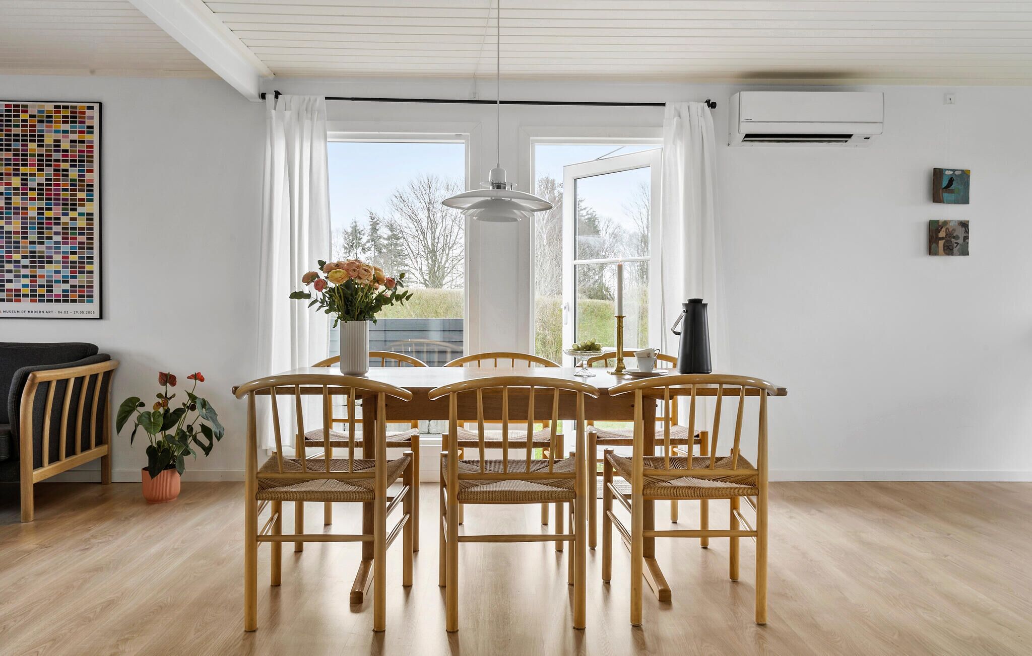 Un'area pranzo con tavolo e sedie in legno, un vaso di fiori e vista sull'esterno tramite ampie finestre.