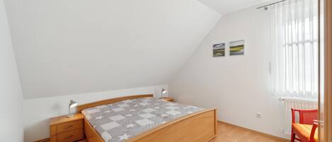 A bedroom with a wooden bed, a nightstand, a desk, a chair, and a window with curtains.