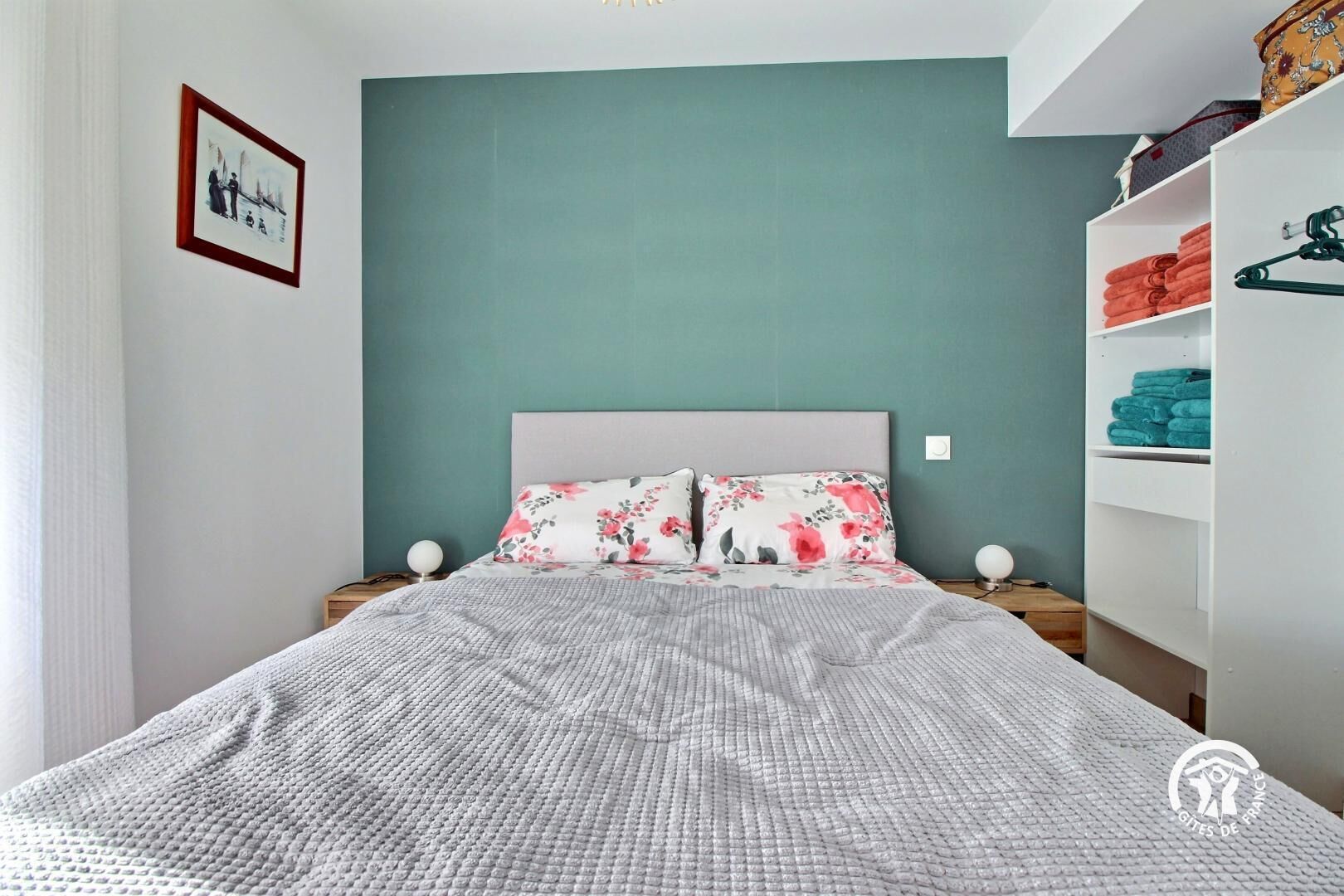 Une chambre avec un mur de couleur turquoise, un lit recouvert d’une couette à motifs floraux et une table de chevet avec une lampe.