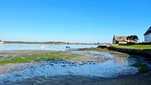 Une scène côtière avec un ciel dégagé, une plage de sable et une étendue d’eau. On aperçoit des bateaux sur l’eau, et un bâtiment avec un toit en pente sur le côté droit.