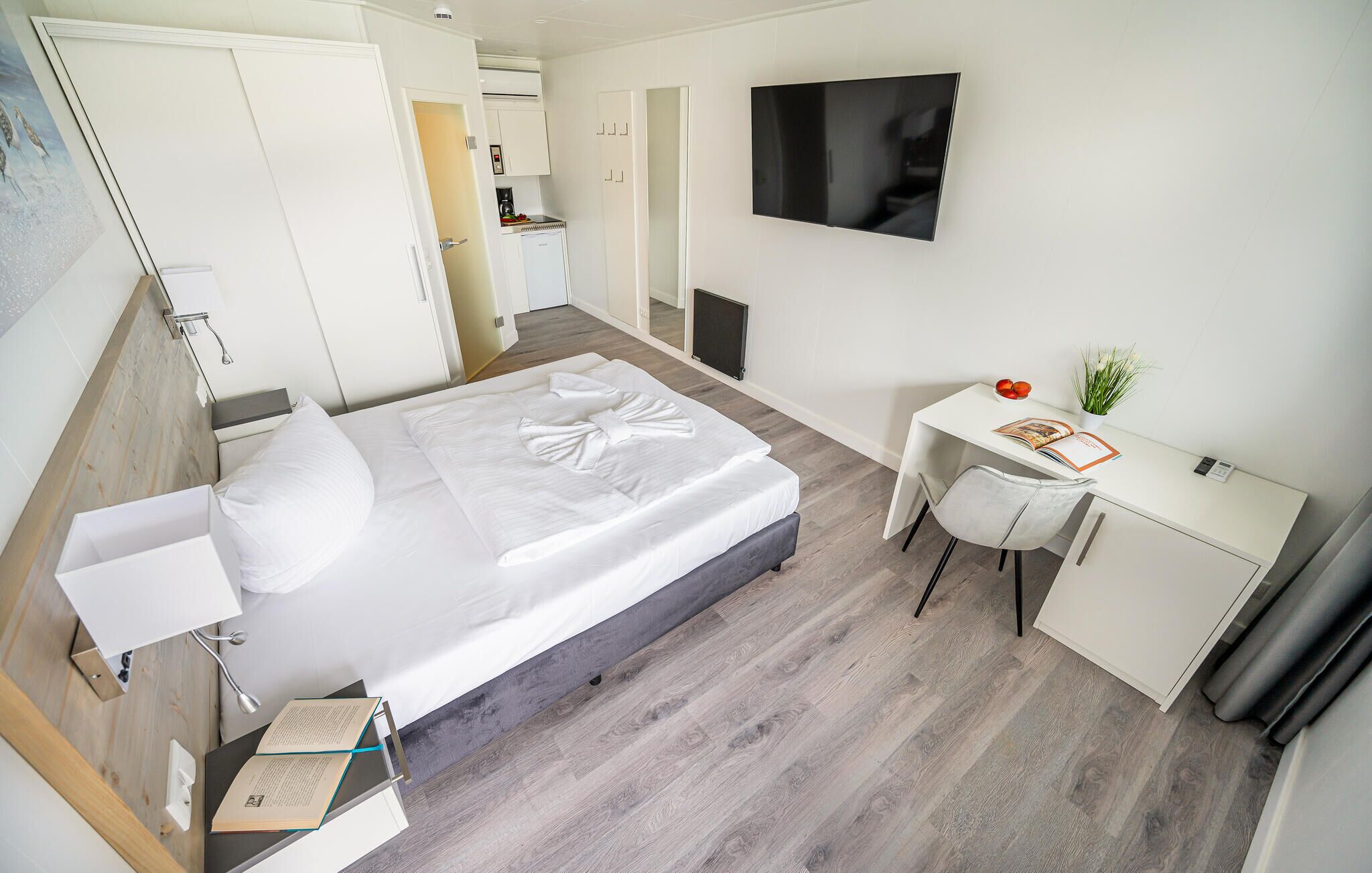 Ein modernes Hotelzimmer mit einem ordentlich bezogenen Bett, einem Schreibtisch mit Stuhl, einem Flachbildfernseher und einer kleinen Küchenzeile.