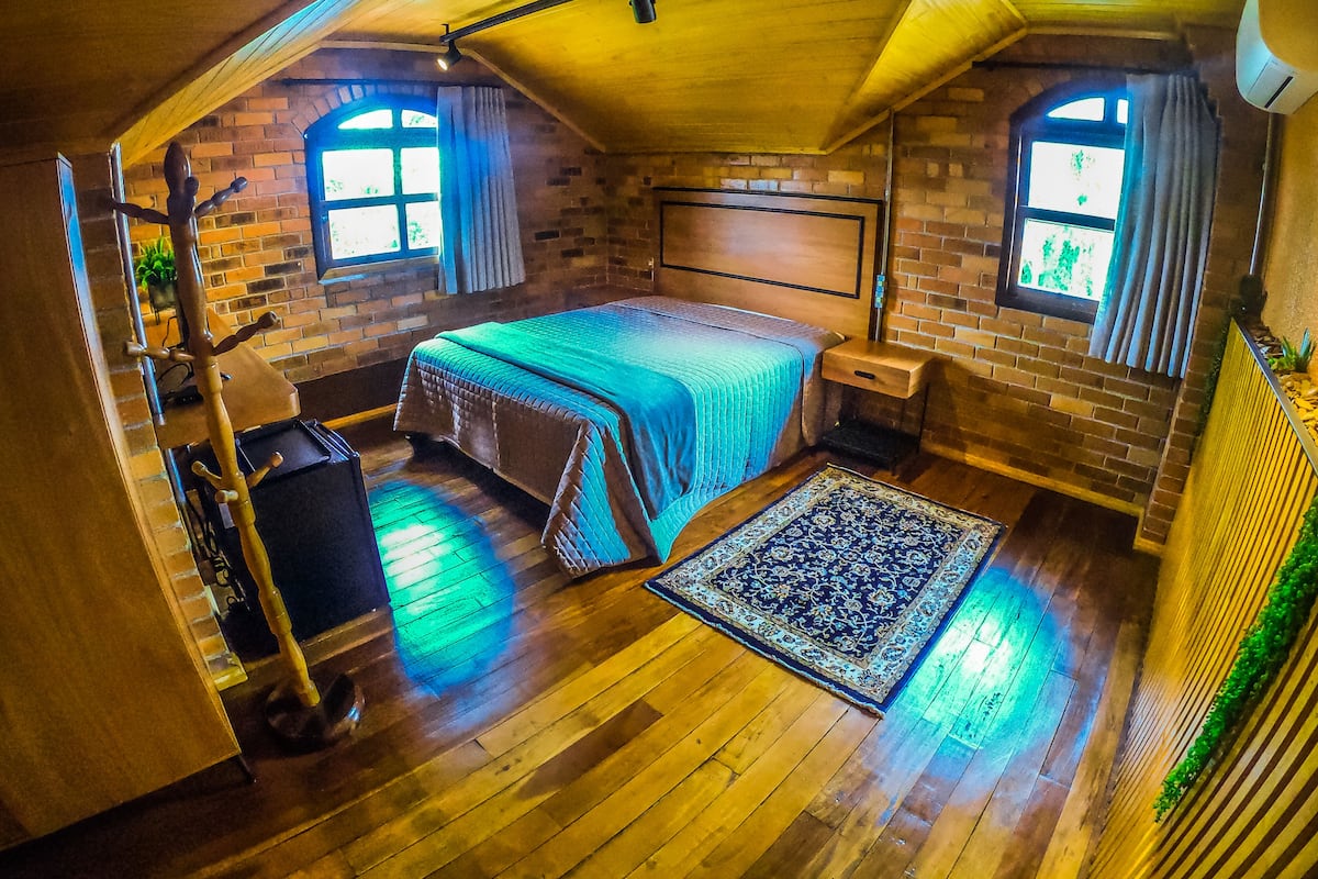 A cozy attic bedroom with a wooden ceiling, brick walls, a bed with a blue bedspread, a nightstand, a dresser, and a patterned rug.