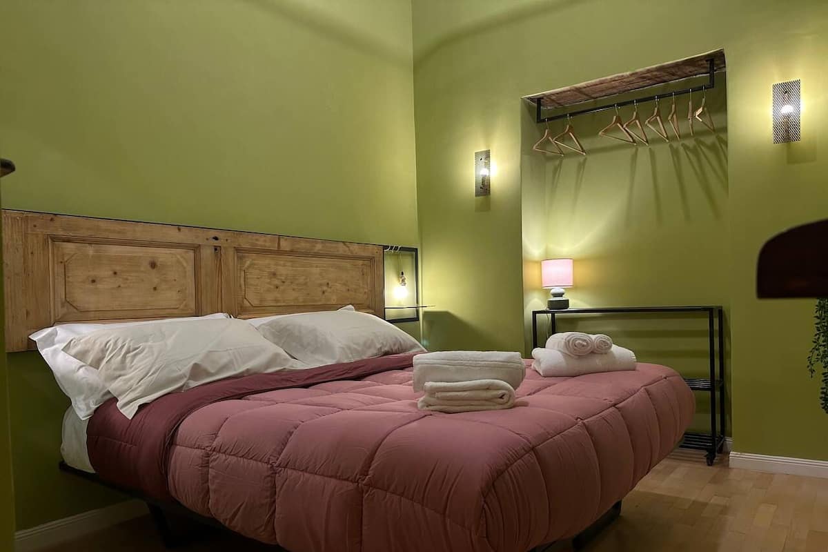 Una cama bien hecha con edredón rosa y almohadas blancas, un cabecero de madera, un toallero en la pared y una mesita de noche con una lámpara.