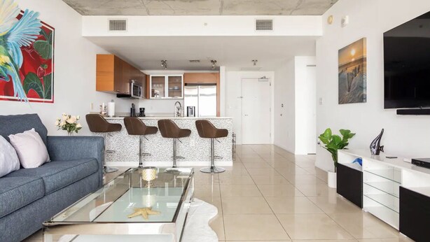 A modern living room with a glass coffee table, a grey sofa, and a kitchen area in the background.
