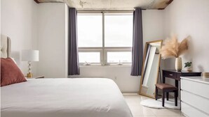 A bedroom with a large bed, a desk, a chair, a dresser, a window with curtains, and a mirror.