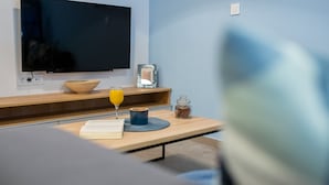Una habitación moderna con una televisión de pantalla plana en la pared, una mesita de madera con un tazón, un vaso de jugo de naranja, un libro y un frasco pequeño.