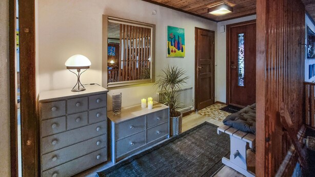 A cozy entryway with a wooden floor, a grey dresser with drawers, a lamp, a potted plant, and a door with a stained glass window. - Nice home in Romelanda with sauna (Romelanda)