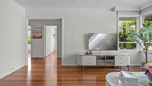 A modern living room with a wooden floor, a white console table, a flat-screen TV, and a large window with a view of greenery. - StayAU 2BRM Beach Family House Hampton (Hampton)