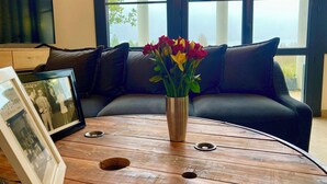 Ein Holztisch mit Blumen in einer Vase, ein gerahmtes Foto und ein schwarzes Ledersofa in einem Raum mit großen Fenstern.