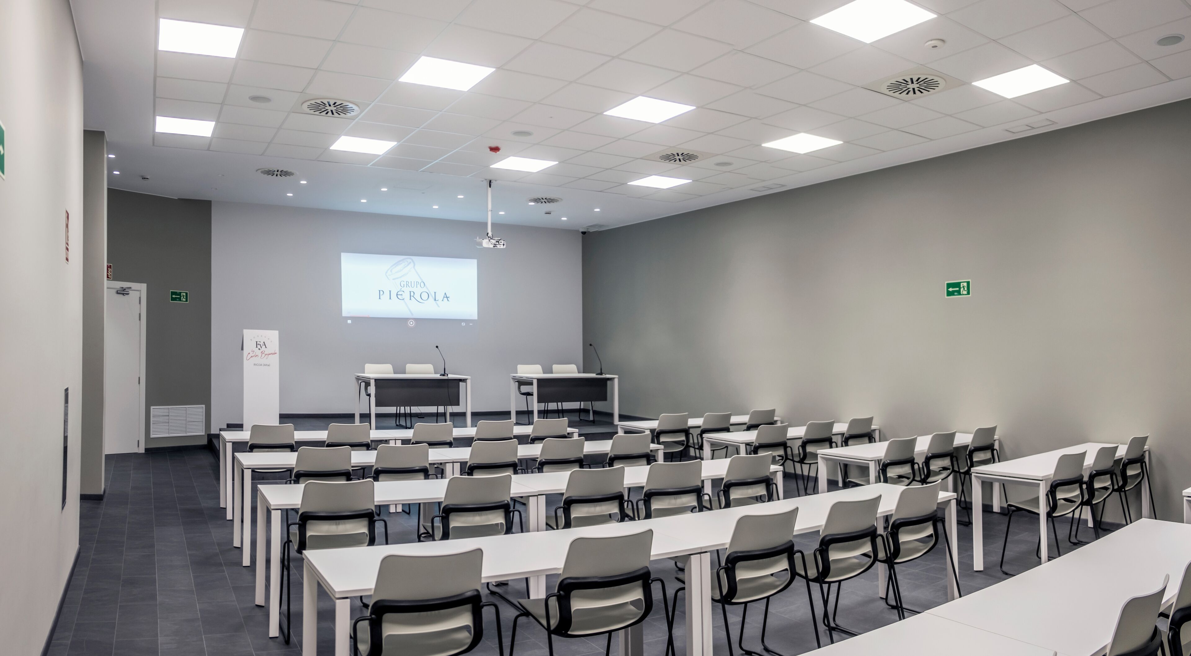 Un salón de conferencias con varias mesas y sillas, una pantalla de proyector mostrando 'Grupo Pierola' y una pizarra.