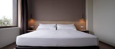A modern hotel room with a large bed, two bedside tables, and a view of greenery outside.