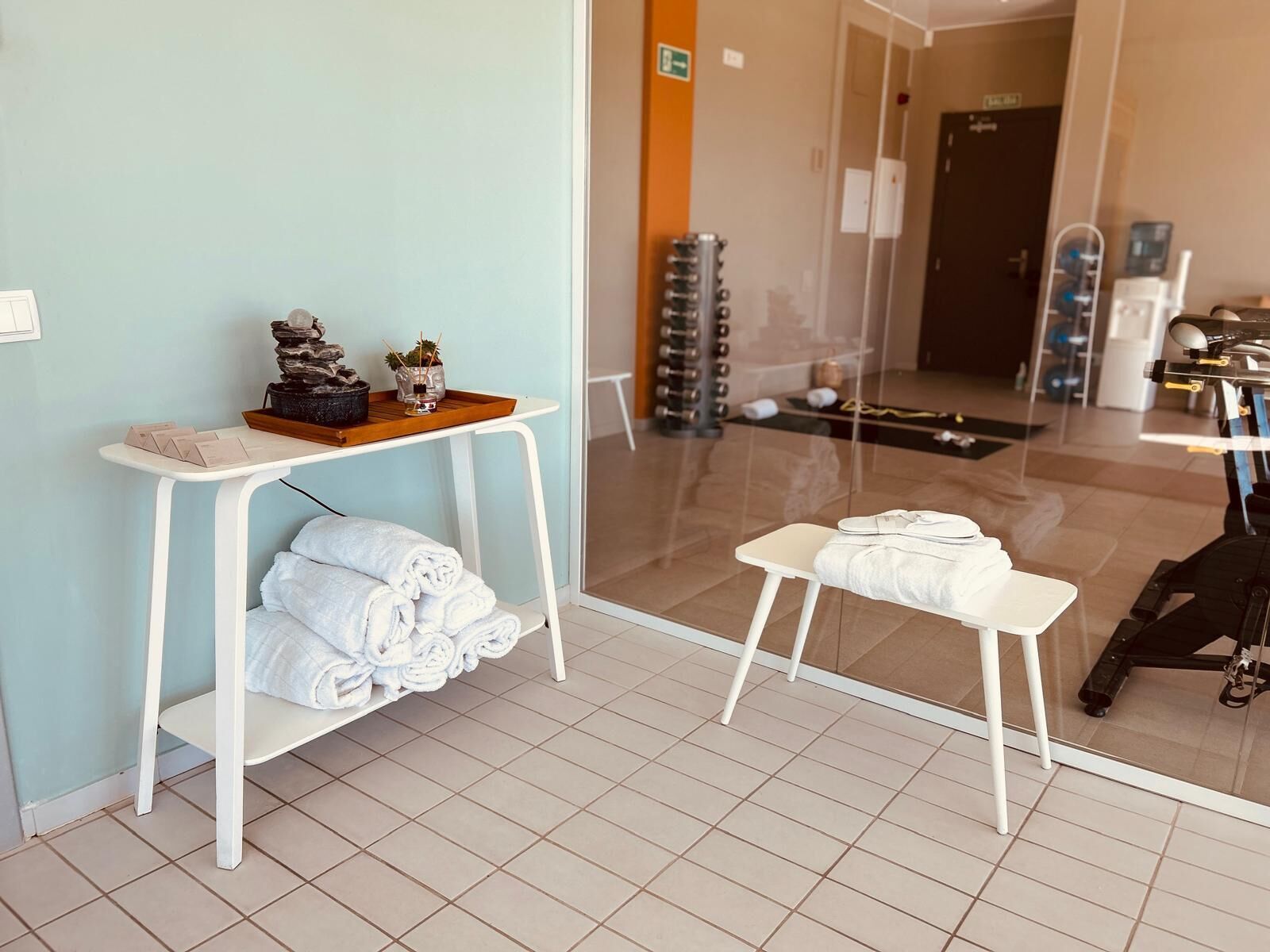 Habitación de hotel con puerta de vidrio que da a un gimnasio, una mesita blanca con toallas y una banca.