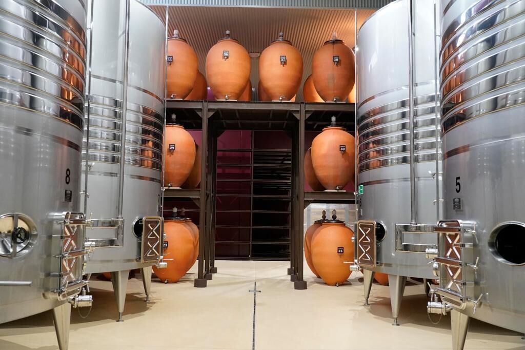 Zona de almacenamiento con tanques metálicos grandes y recipientes cerámicos más pequeños, probablemente para vino o elaboración de cerveza.