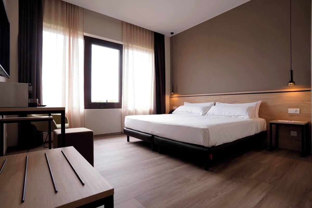 A hotel room with a bed, a desk, a chair, a lamp, and a window with curtains.