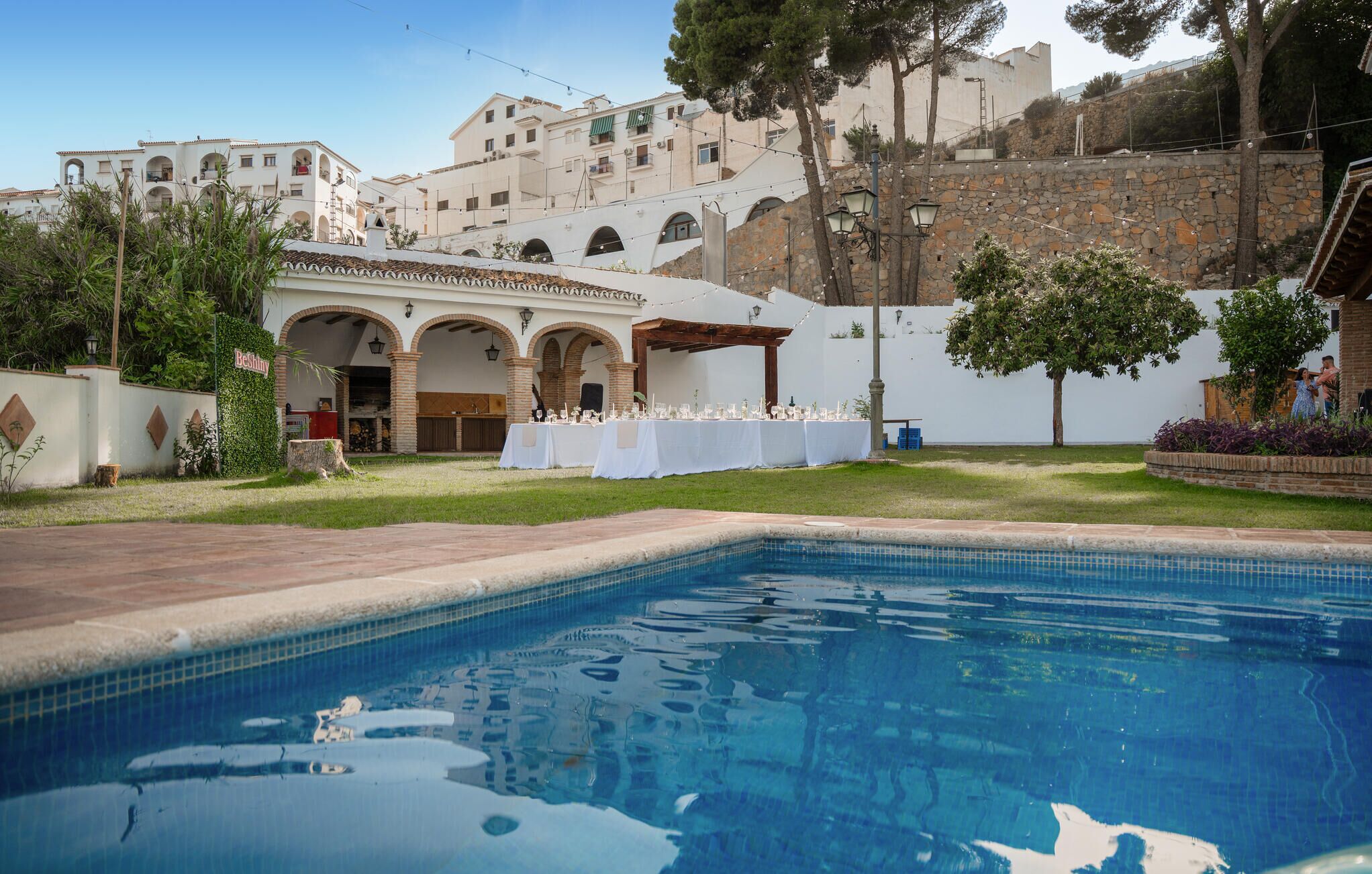 Zona de piscina con mobiliario de comedor, pared de piedra y un edificio con arcos al fondo.