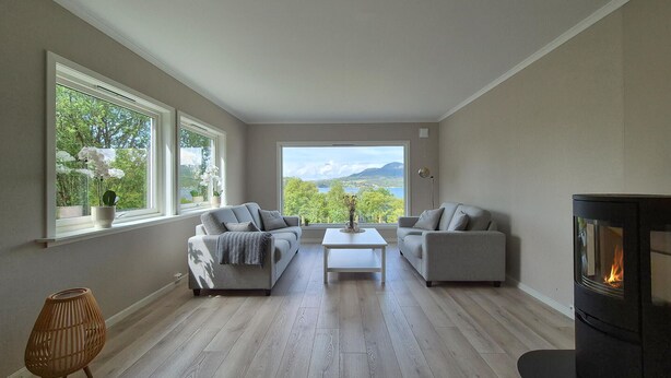 A living room with two grey sofas, a white coffee table, a wicker floor lamp, and a fireplace. Large windows offer a view of greenery and mountains. - Nice home in Vikebygd with house sea view (Vikebygd)