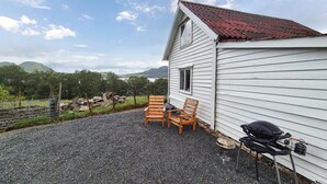 A white building with a sloped roof, a gravel patio, two wooden chairs, and a barbecue grill. - Nice home in Vikebygd with house sea view (Vikebygd)