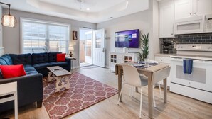 Un salon moderne avec un canapé, une table basse et un téléviseur. Il y a également un coin repas avec une table et des chaises. La cuisine est équipée de meubles blancs et d’un four à micro-ondes.