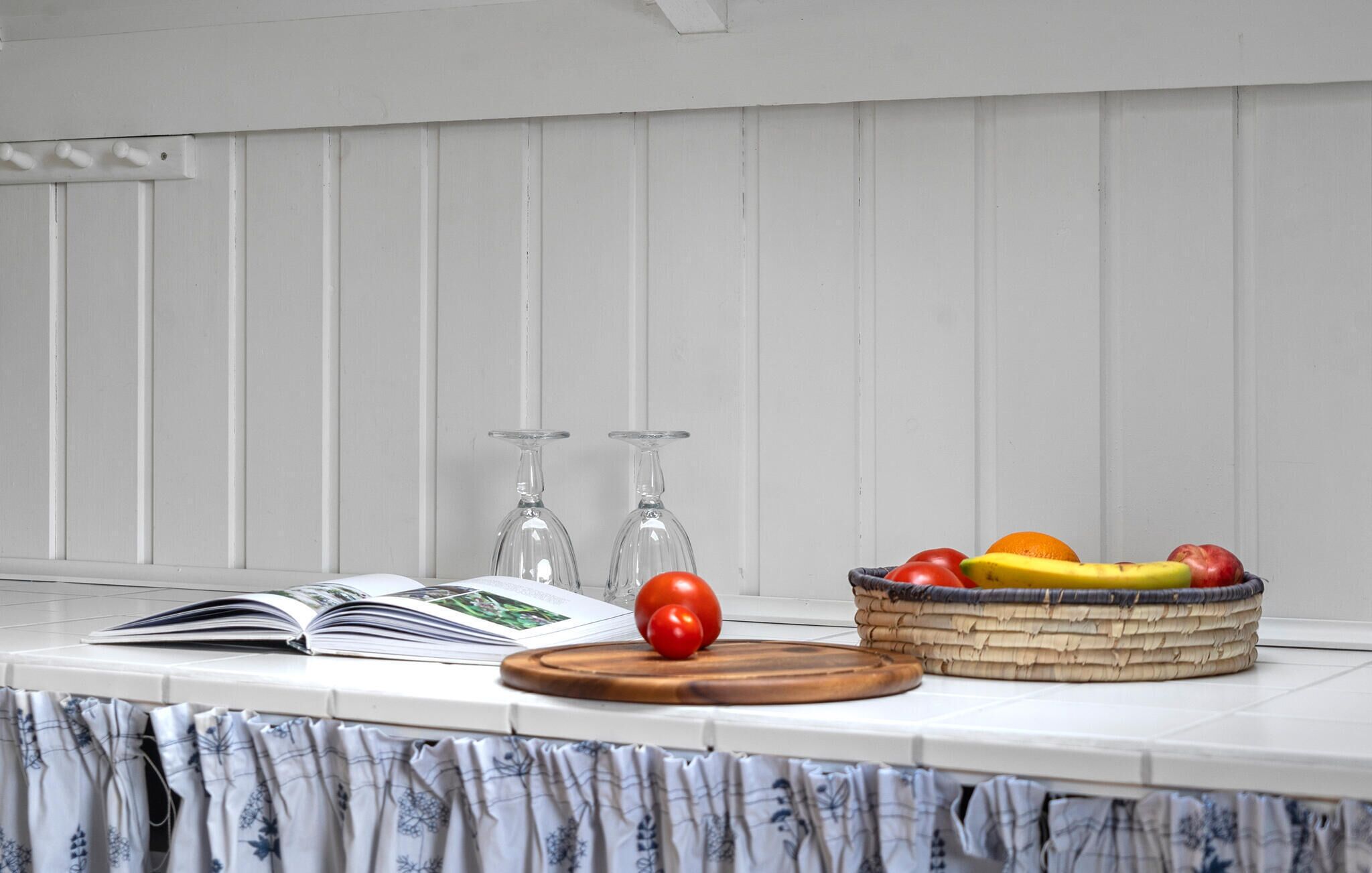 A table with a book, two wine glasses, a wooden cutting board, and a basket of fruit.