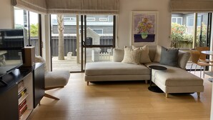 A modern living room with a sofa, a coffee table, and a television. There is a sliding glass door leading to a balcony with a view of the outside.