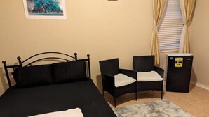 Une pièce comprenant un lit, deux chaises, une petite table, un réfrigérateur et un tableau accroché au mur.