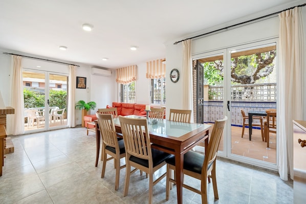 A dining area with a glass-top table and wooden chairs, a red sofa, and a view through a glass door to an outdoor patio.